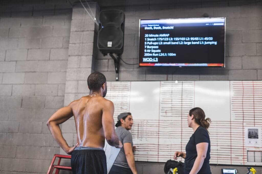 Gym members by a whiteboard under a wall-mounted screen showing the workout of the day