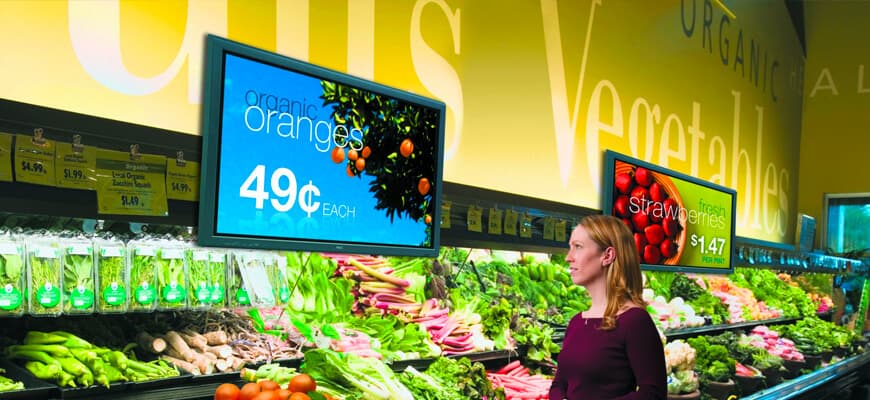 A shopper in a grocery produce aisle looking at two screens with fruit prices