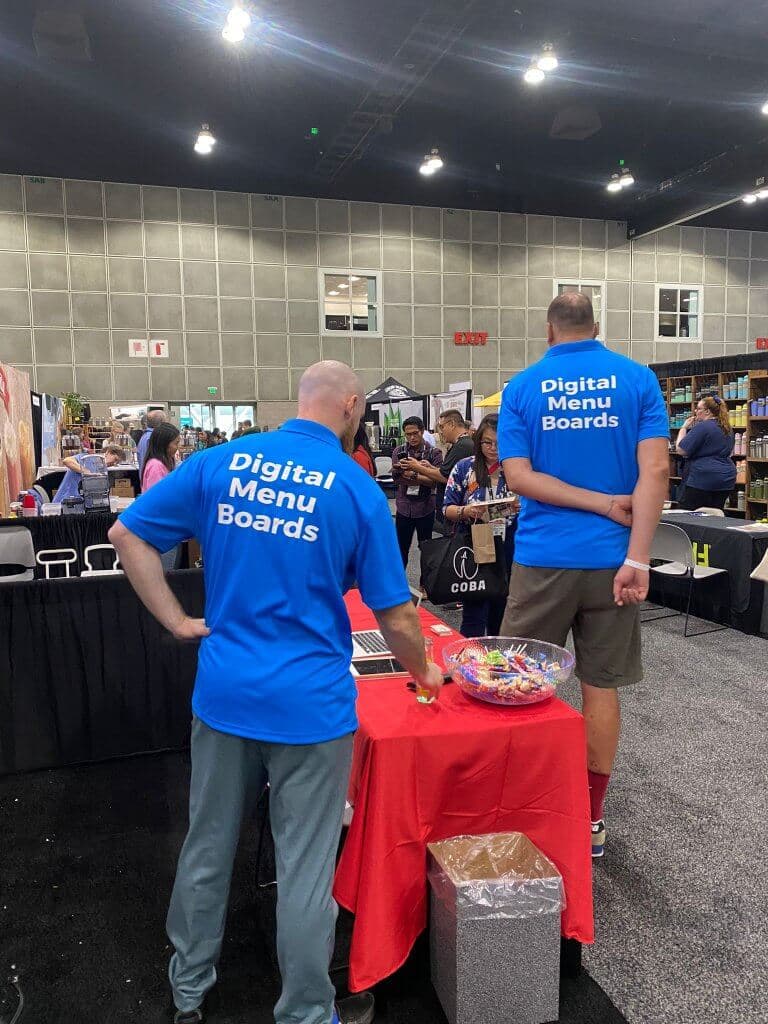Two Liqvid team members seen from behind in shirts reading Digital Menu Boards, at the booth in the exhibition hall